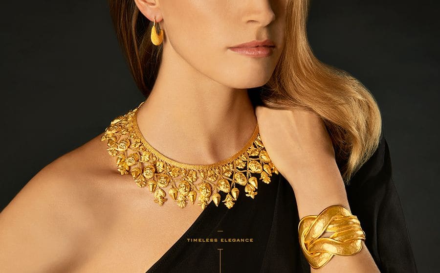 Woman wearing an elegant gold necklace, matching gold bracelet, and drop earrings, styled with a black off-shoulder outfit.